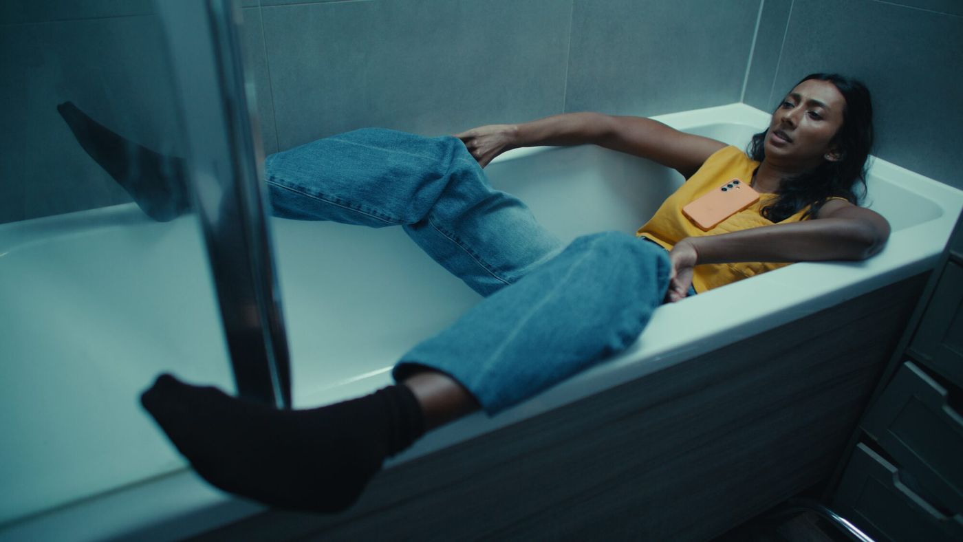 A woman lies fully clothed in an empty bath, phone in hand, lit cool blue