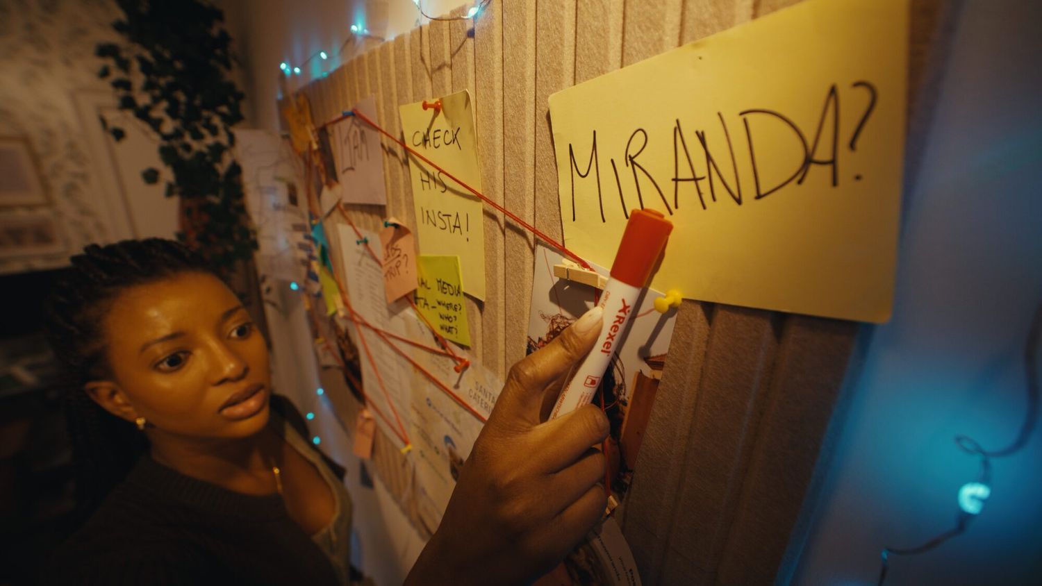 A woman pins a note reading Miranda to a corkboard strung with red thread