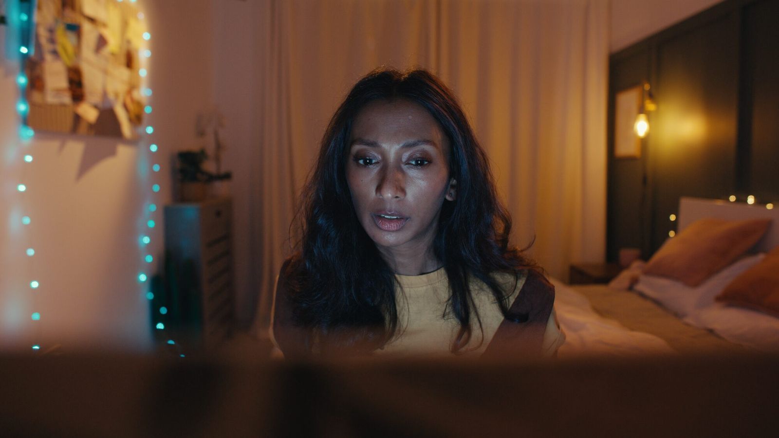 A woman sits up in bed at night, lit by a laptop screen, fairy lights behind her