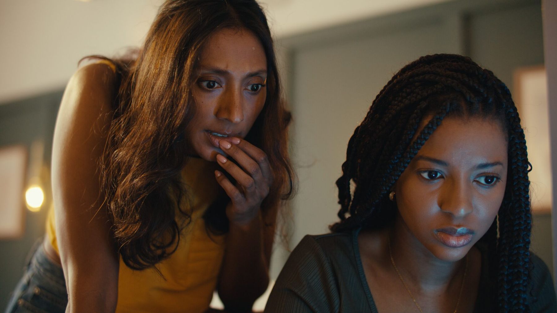 Two women lean over a laptop in a warmly lit bedroom, faces caught in its glow